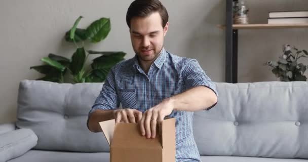 Молодий красивий усміхнений чоловік розпаковує маленьку картонну коробку . — стокове відео