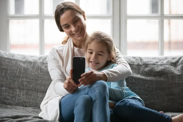 Smiling mom and small daughter use smartphone at home