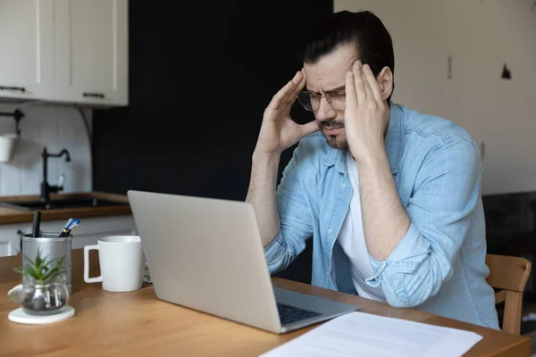 Přepracovaný mladý muž trpící bolestí hlavy v domácí kanceláři. — Stock fotografie