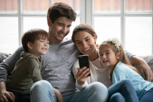 Happy parents relax on couch using cell with kids