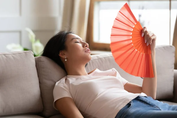 Overheated exhausted Asian woman waving paper fan, sitting on couch — Stock Photo, Image