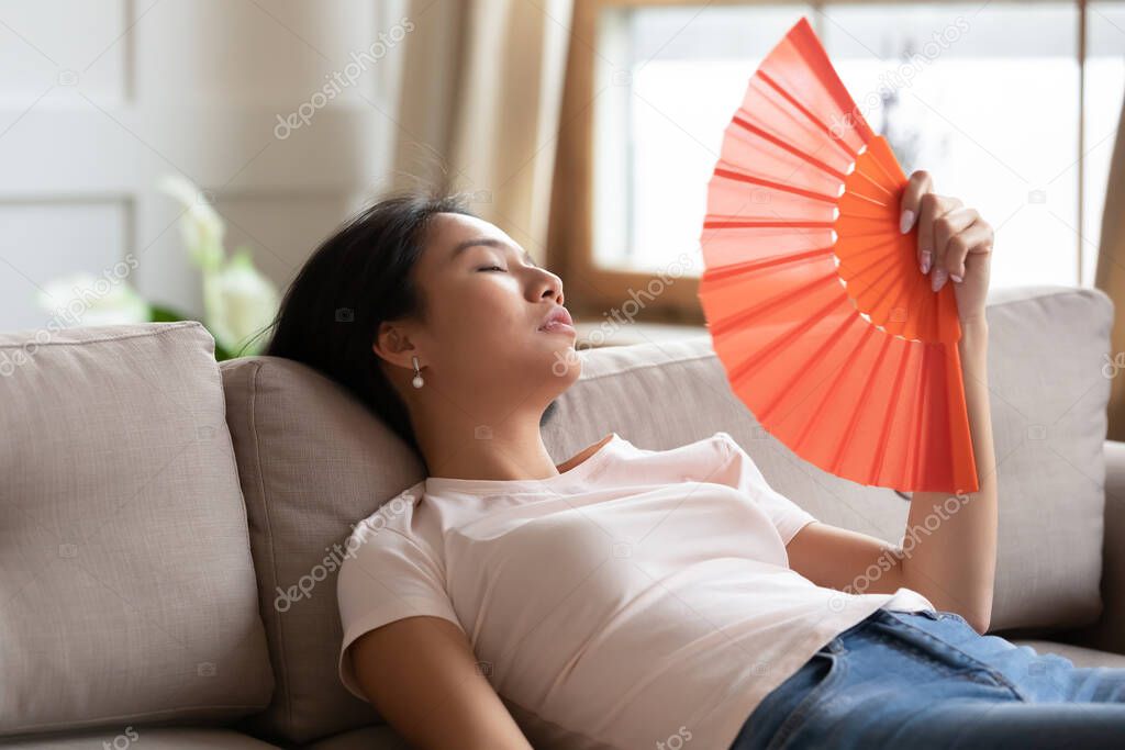 Overheated exhausted Asian woman waving paper fan, sitting on couch