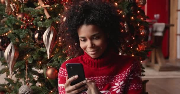Mujer africana usando teléfono inteligente relajante sentado cerca del árbol de Navidad decorado — Vídeo de stock