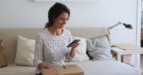 Счастливая женщина сидит на диване, держа смартфон открывает коробку с посылками — стоковое видео
