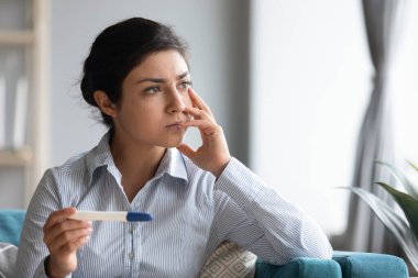 Elinde hamilelik testi olan üzgün Hintli kadın pencereden dışarı bakıyor.