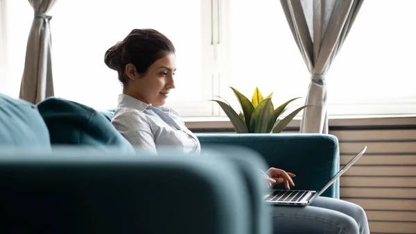 Indian female sit on couch working on laptop from home