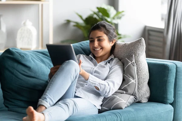 Relaxado indiana menina deitada no sofá segurando tablet desfrutar de e-shopping — Fotografia de Stock