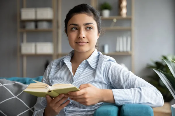 Chica india sentada en el sofá con libro mirando a la distancia — Foto de Stock