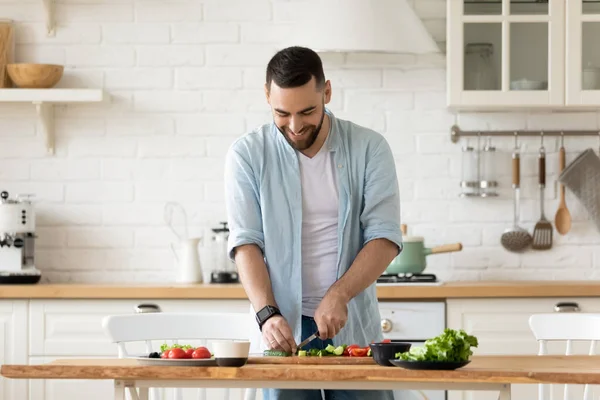 Gülümseyen genç adam salata pişiriyor, modern mutfakta sebze kesiyor. — Stok fotoğraf