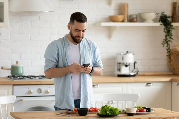 Genç adam akıllı telefon kullanıyor, mutfakta salata pişiriyor, yemek tarifi arıyor. — Stok fotoğraf