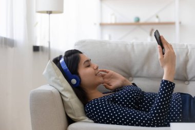 Genç Asyalı kadın kanepeye uzanıp müzik dinliyor.