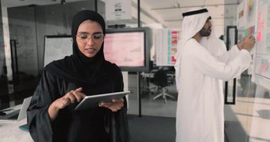 Kafiyeli genç Müslüman iş kadını ofiste tablet kullanıyor. Arkalarından konuşan, online proje üzerinde çalışan, internet iletişimi için internet teknolojisini kullanan Arap erkek arkadaşları var.