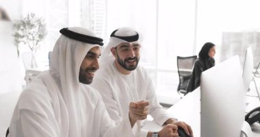 Beyaz Müslüman elbiseli iki Arap erkek iş arkadaşı masaüstü monitöründe online video ürününü tartışıyor, birlikte oturuyorlar, içeriği izliyorlar, gülümsüyorlar, gülüyorlar.