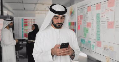 Beyaz Müslüman elbiseli, ciddi genç Arap profesyonel, arka planda meslektaşlarıyla birlikte akıllı telefonda yazı yazıyor, cep telefonunda proje yönetimi için uygulama kullanıyor.