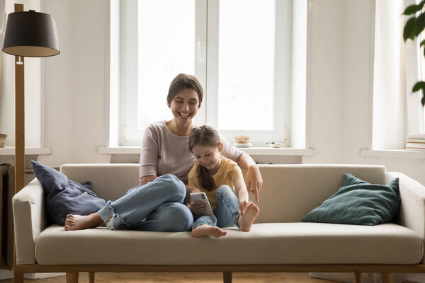 Screen time with love. Cheerful laughing mother small daughter rest on couch at cozy family home cuddle hold cellphone enjoy online shopping play face filters game app read funny messages from chat