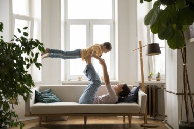 Hayali uçuş. Neşeli aktif genç anne kanepede sırt üstü uzanıyor. Havada küçük tatlı bir kız eğik bacakları üzerinde. Heyecanlı çocuk jimnastik egzersizleriyle eğlenceli bir uçak oyunu oynuyor.
