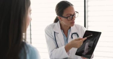 Gözlüklü ve beyaz önlüklü genç beyaz kadın travma cerrahı hasta kadına röntgen görüntüsü gösteriyor, gülümsüyor, gülüyor, muayenehanede danışmanlık yapıyor.