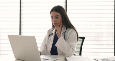 Bilgisayardaki elektronik tıbbi raporlar üzerinde çalışan, klinikteki çalışma masasında oturan, dizüstü bilgisayarda yazan, çeneye dokunan, düşünen, gülümseyen, göz gezdiren genç doktor kadın.