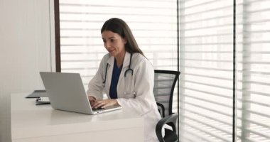 Genç Latin pratisyen kadın ofisten bir hastayla sohbet ediyor, çalışma masasında dizüstü bilgisayarla yazı yazıyor, başka tarafa bakıyor, internet iletişimini düşünüyor, online danışmanlık yapıyor.