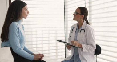 Ciddi genç kadın pratisyen kadın gözlüklü ve laboratuvar önlüklü. Muayeneden sonra kanepede oturan hasta kadına danışmanlık yapıyor.