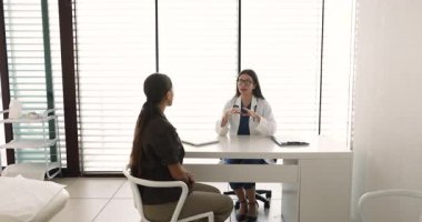 Kadın doktor ve hasta muayenehanede buluşuyor. Beyaz iç mekan, çalışma masasında konuşuyor. Pratisyen kadın terapi için müşteriye danışmanlık yapıyor, iyileşme süreci başarılı.