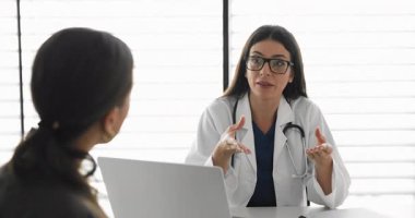 Gözlüklü ve laboratuvar önlüklü kendine güvenen kadın doktor hastayı dinliyor, konuşuyor, başarılı terapi, tedavi, rehabilitasyon için tavsiyelerde bulunuyor, tıp bürosunda bilgisayarla aynı masada oturuyor.