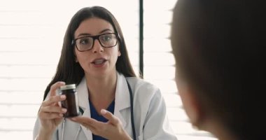 Gözlüklü, beyaz önlüklü, kadın hastaya ilaç sunan, ilaç, vitamin, gıda takviyesi yapan, elinde cam şişe tutan kendine güvenen genç Latin bir kadın.