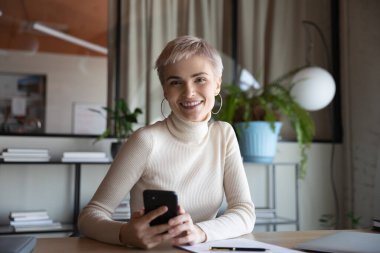 Ofis portresindeki akıllı telefonlu mutlu kısa saçlı iş kadını. Çalışan, girişimci, cep telefonu tutan lider masada oturuyor, dişlek gülümsemeyle kameraya bakıyor. Kafasından vur.