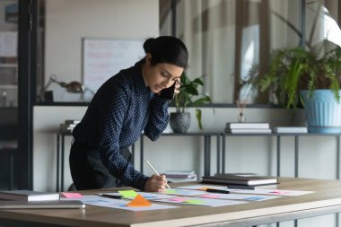 Birçok işi olan Hintli genç bir iş kadını yoğun bir şekilde çalışıyor. Yapışkan kağıtlar, belgeler, grafik raporları, not yazma, cep telefonuyla konuşma, cep telefonuyla konuşma.