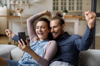 Heyecanlı genç evli çift güzel sürpriz haberlerle kısa mesaj alıyor, cep telefonu ekranına bakıyor, neşe için gülüyor, kazanan el hareketi yapıyor, galibiyeti kutluyor, başarı,