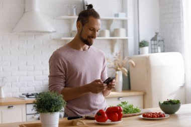 Pozitif genç ev sahibi mutfakta akıllı telefon kullanıyor, salata tarifi okuyor, sağlıklı yemek pişiriyor, kız arkadaşıyla akşam yemeği için taze sebze salatası pişiriyor, internette sohbet ediyor, mesaj yazıyor.