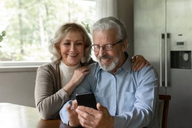 Cihazı birlikte kullanmak. Meraklı yaşlı eş, sevgili kocanın omuzlarını kucaklıyor ve onu mobil web uygulamasında oynarken izliyor. Mutlu yaşlı evli çift cep telefonuyla boş vakit geçirir alışveriş haberleri yapar.