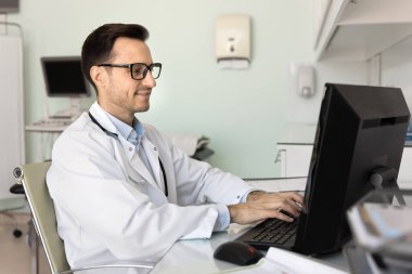 Doktorların iş günlerinde evrak işleri. Klinikte çalışan orta yaşlı, kendine güvenen bir erkek sağlık uzmanı not yazmak için bilgisayarını kullanır. Tıbbi veri yazar, hastalara danıştıktan sonra sisteme reçete yazar.