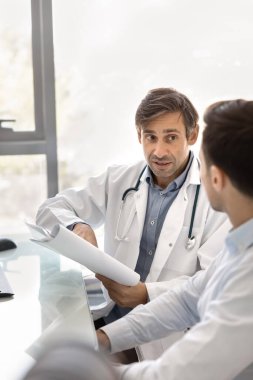 İlaç konsültasyonu. İki orta yaşlı adam doktor ilaç temsilcisi tıbbi dosyaları inceleyip klinikteki masada belgeleri tartışıyorlar. Doktor, tedavi veya ürün detaylarını temsilciye açıkladı