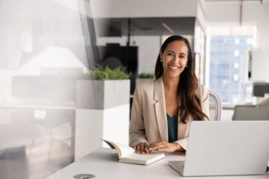 Neşeli genç Yeni Zelandalı proje yöneticisi iş yerinde dizüstü bilgisayarı açık bir ofiste oturan dişlek gülümsemeli, gülen, profesyonel portre için poz veren bir kadın.