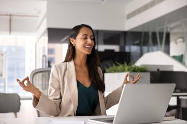 Mutlu ve dingin genç Yeni Zelandalı iş kadını iş yerinde sakin duruyor, zen parmağı hareketleriyle oturuyor ve gözlerini kapıyor, gülümsüyor, ofis alanında meditasyon yapıyor, ahenk kuruyor, dengeyi kuruyor.