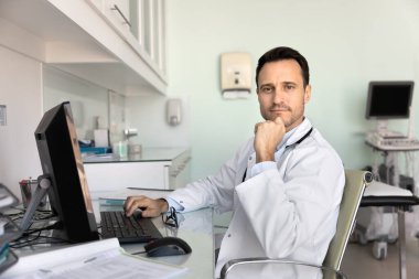 Kendine güvenen bir doktor. Portre, orta yaşlı, orta yaşlı, erkek, sağlık uzmanı. Kameraya bakarken dikkati dağılıyor. Masaüstü bilgisayarında kayıt yapıyor.