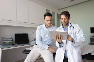 Görsel sağlık eğitimi. Laboratuvar önlüklü profesyonel erkek doktor GP kardiyoloğu konsültasyondaki hastayla konuşuyor. Tablet ekranındaki dijital görüntü tanı verileri hastalık tedavisini açıklıyor.