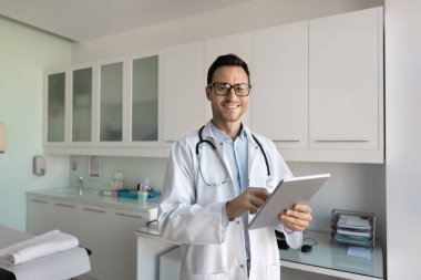 Modern hastane çalışanı. Ofiste poz veren mutlu arkadaş canlısı erkek doktor kameralı tutucu tabletine bakıyor ve tele-sağlık uygulaması kullanarak hastaya uzaktan danışmaya hazır. Boşluğu kopyala