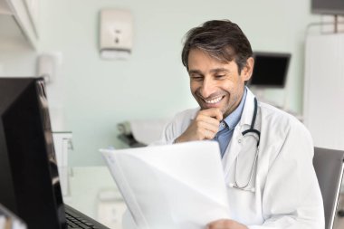 Gülümseyerek kağıtları kontrol et. Klinikte evrak işleriyle uğraşan mutlu erkek doktor basılı fotokopi inceleme hasta kurtarma raporundan heyecan verici araştırmalar okumaktan gurur duyar.