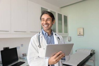 Tabletli neşeli bir doktor. Mutlu gülümseyen erkek doktor klinikte bilimsel başarılardan tatmin olmuş dijital tabaka kullanarak duruyor. Teşhis ameliyatı, tele sağlık uygulamasında kolay bir çalışma sağlıyor.