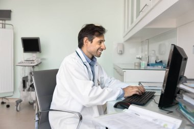 Sağlık hizmetlerinde idari iş verimliliği. Gülümseyen erkek doktor klinisyeni iş yerinde oturup sigorta evrakları hazırlıyor başarılı hasta vakalarıyla ilgili bilimsel makaleler yazıyor.