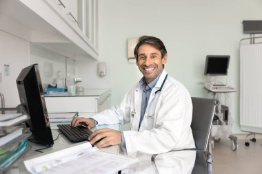İş yerinde tıbbi uzman. Portre, olgun erkek doktor. Masada kağıtlar ve bilgisayarla oturmuş kameraya bakıyor. Başarılı sağlık uygulamalarının tadını çıkarın.