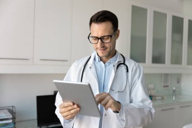 Doktor bağlantıda kalıyor. Tıbbi sohbet yoluyla hasta ile iletişim kuran, beyaz önlük takan gülümseyen, deneyimli erkek doktor klinikteki tablet kullanarak reçete kayıtları yapıyor.