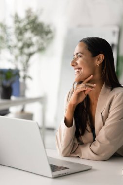 Neşeli genç Yeni Zelandalı iş kadını şirket büyümesi, proje başarısı, başarılı kariyer ofiste dizüstü bilgisayarda oturan, çeneye dokunan, yüzünü çeviren, gülümseyen. Dikey çekim