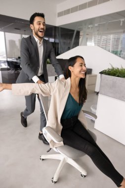 Sandalyeye oturan neşeli genç ofis çalışanları. Neşeli mutlu erkek çalışan heyecanlı bayan meslektaşıyla koltuğu ileri itiyor, gülüyor, eğleniyor, iş başarısını kutluyor.