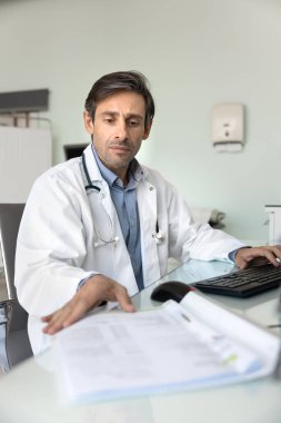 Doktor evrak işlerini hallediyor. Masa başında, kağıt dosyalardaki laboratuvar sonuçlarıyla odaklanmış yetişkin erkek tıbbi uzmanlık mesleği. Hasta verilerini çevrimiçi e-sağlık platformu inceleme taleplerine girin.