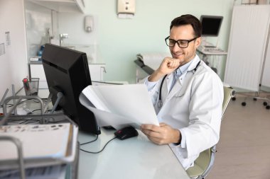 Rutin idari işler. Gülümseyen erkek doktor klinik ofisindeki bilgisayarın yanında oturuyor. Hasta ilerleme raporunun yeni araştırma verileriyle birlikte incelendiğini görmekten memnuniyet duyuyorum.