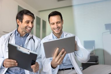 Sağlık hizmetlerinde dostça iletişim. İki neşeli erkek doktor rahatlayıp, samimi kahkahalar paylaşıyor. Tablet bilgisayar ekranında dijital kayıtlar. Günlük sohbetin tadını çıkarıyorlar. Boşluğu kopyala