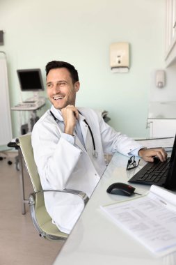 Sağlık hizmetleri gününde neşeli bir an. Gülen erkek doktor hastane masasına oturur arkana bakar rahatla iş arkadaşının dijital belgeleri incelemekten dikkati dağılan şakasının keyfini çıkar bilgisayardaki bilimsel evraklar.